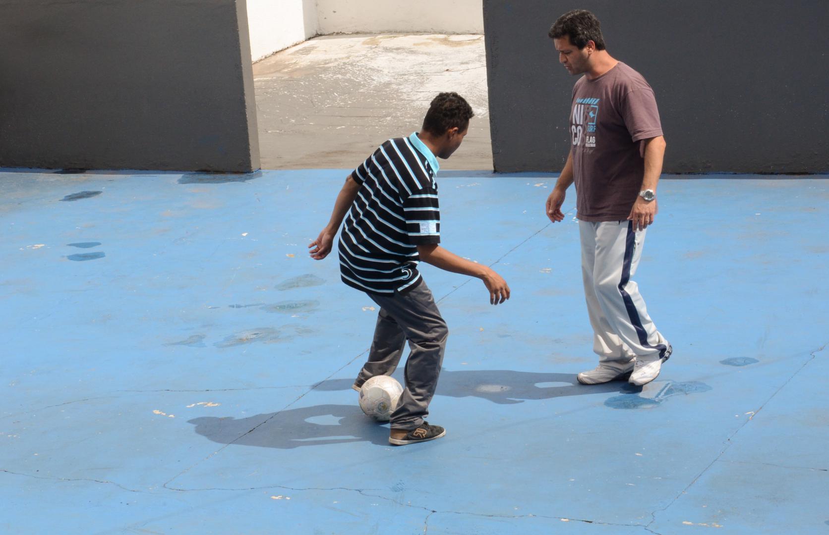 a imagem mostra professor e aluno na quadra de esportes, em pé, frente a frente. O professor olha e fala com o aluno, que está com a bola, na cor branca, entre os pés, fazendo movimento de futebol. O aluno está de costas para a foto e o professor, de frente. A quadra tem chão na cor azul.a imagem mostra professor e aluno na quadra de esportes, em pé, frente a frente. O professor olha e fala com o aluno, que está com a bola, na cor branca, entre os pés, fazendo movimento de futebol. O aluno está de costas para a foto e o professor, de frente. A quadra tem chão na cor azul.