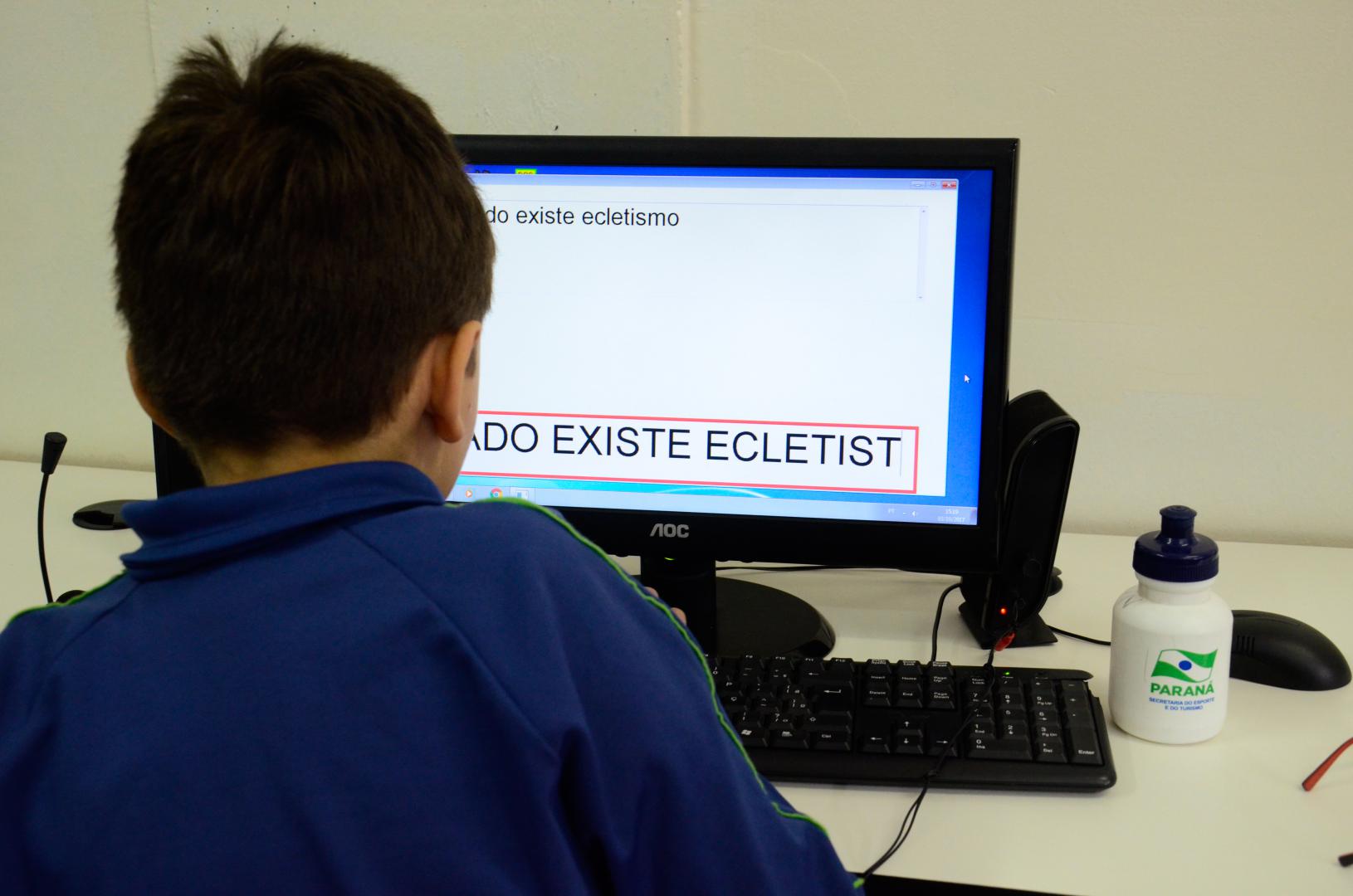 a imagem mostra um aluno criança de costas, sentado de frente para um computador, que tem texto sendo mostrado na tela.