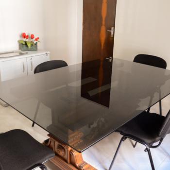 a foto mostra uma das salas de reunio, com uma mesa com tampo preto retangular e quadro cadeiras pretas ao redor dela. Ela tem um p em madeira marrom todo trabalhado. Ao fundo est um pequeno armrio branco, baixo, de duas portas, com uma bandeja e um vaso de flores vermelhas sobre ele. Ao fundo e  direita est uma porta em madeira marrom, fechada. As paredes e o cho so brancos.