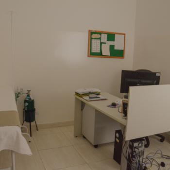 a foto mostra a enfermaria, com uma mesa com computador à direita, branca. Há alguns papéis sobre ela. Ao fundo, há um mural de recados na cor verde. À esquerda está a cama para exames. As paredes e o chão são brancos.