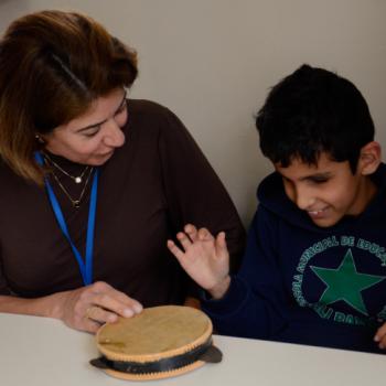 a imagem mostra professora e aluno sentados a uma mesa branca, lado a lado, sendo ela  esquerda da foto. Sobre a mesa h um pequeno pandeiro branco, com couro na cor marrom clara. A professora tem a mo direita sobre o pandeiro. Ela olha para o aluno,  sua esquerda ( direita da foto) e sorri, com a cabea levemente tombada para o lado. O aluno tambm sorri. Ele tem a cabea baixa, olhando para o pandeiro, e a mo direita erguida como fazendo movimento para bater no instrumento musical.