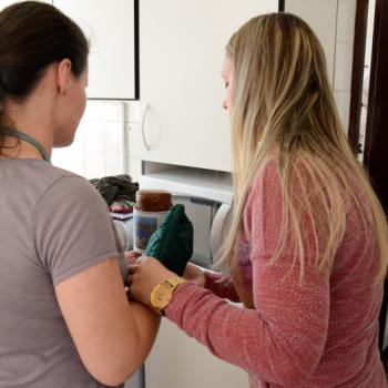 a imagem mostra aluna  esquerda da foto e professora  direita, ambas de costas para a imagem. Elas esto na cozinha. A professora ajuda a aluna a colocar na mo direita uma luva trmica na cor verde escuro, dessas utilizadas para retirar recipientes quentes do forno. A frente delas,  possvel ver parte de um microondas e de um armrio, ambos na cor branca.