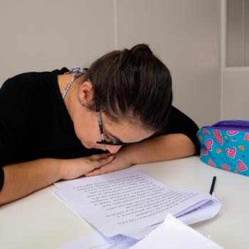 : a imagem mostra uma aluna adolescente sentada  mesa branca, lendo texto em papel branco sobre a mesa. Ela olha o papel bem de perto. Usa culos. Tem os dois braos, at o cotovelo, abertos sobre a mesa, sendo a mo direita sobre a esquerda. Ao seu lado esquerdo (direito da foto), h um penal na cor azul com coraes coloridos e de tamanhos variados.