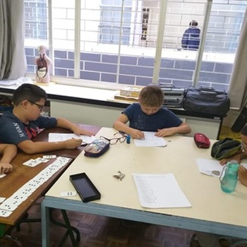 Foto de quatro meninos envolta de duas mesas. Eles escrevem em papeis.