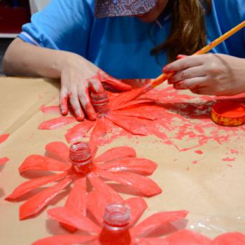 a imagem mostra as mos de uma aluna pintando de vermelho garrafas pet cortadas em formato de flores. Ela est sentada a uma mesa coberta com papelo marrom. Tem as mos sujas de tinta. A mo direita est sobre a mesa, com o dedo indicador dentro de um pequeno pote de tinta. A mo esquerda segura um pincel sobre uma das ptalas.  frente da estudante h trs flores j pintadas de vermelho.