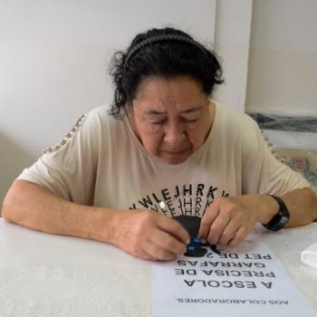 a imagem mostra uma senhora sentada a uma mesa branca, fazendo colagem sobre um papel branco com texto. Ela est utilizando as duas mos na atividade, est olhando para baixo, concentrada.