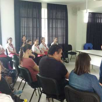 A foto mostra a sala da oficina cheia.  esquerda da imagem esto os participantes sentados, de costas para a foto, concentrados na professora que est em p a frente deles,  direita da foto, falando.