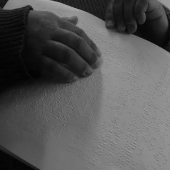 a imagem mostra recorte nas mos de uma pessoa lendo o texto da pea em braille. Ela est tateando o papel branco. A foto  em preto e branco.
