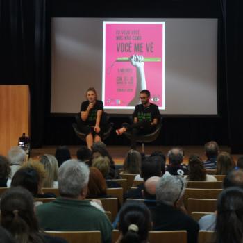 A foto mostra o auditrio de trs pra frente. Tem o pblico sentado de costas e, a frente deles, o palco onde esto sentados uma mulher,  esquerda da foto, e um homem,  esquerda da foto. A mulher segura um microfone e fala. Atrs deles est a projeo do cartaz da campanha.