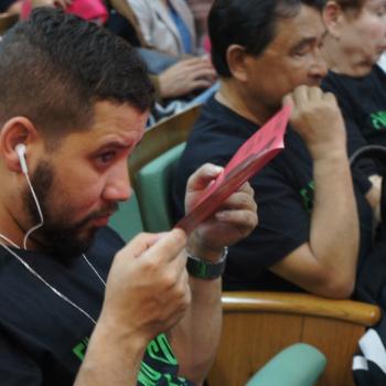 A foto mostra um jovem sentado no auditrio, segurando com as duas mos o panfleto do evento, que l prximo ao rosto. Ele est com fone de ouvido.