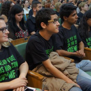 A foto mostra uma fileira do auditrio com pessoas sentadas nas cadeiras. Em plano prximo, est uma mulher, tendo dois jovens ao seu lado esquerdo e uma jovem ao lado deles. Todos usam a camiseta da campanha e olham para a frente. A mulher sorri, os outros tm semblantes leves e meio sorriso.