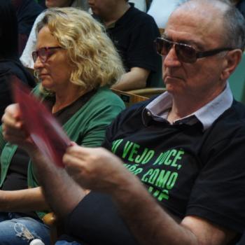 Um homem usando culos e a camiseta da campanha est sentado no auditrio, segurando com as duas mos na altura do peito o panfleto da campanha, que ele l concentrando.