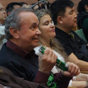 A imagem mostra um senhor sorrindo e olhando para frente, sentado na plateia, segurando com as duas mos a bengala verde dobrada.