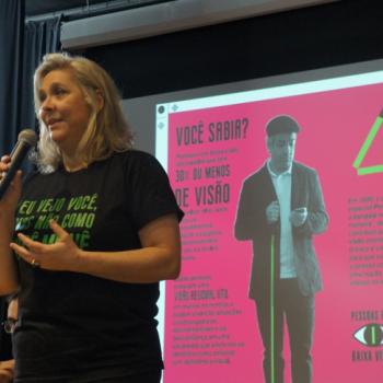 A foto mostra a professora Lilian Biglia, coordenadora da campanha,  esquerda, de lado para a foto, segurando um microfone com a mo direita, gesticulando com a mo direita e falando. Atrs dela, est a projeo em tela do material da campanha.