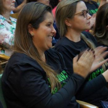 A imagem mostra uma moa sorrindo e aplaudindo, olhando para a frente.