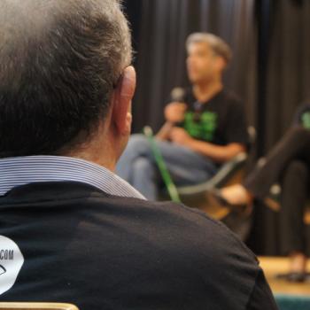 A foto mostra um homem de costas, em primeiro plano, usando a camiseta da campanha que tem nas costas o logo Pessoas com Baixa Viso. Ele olha para frente, onde est o palco com duas pessoas sentadas:  esquerda, Manoel Negraes, colaborador do IPC e coordenador da campanha. Ele segura um microfone e est falando. Ao lado dele est Eliana Cunha, da fundao Dorina Nowill.