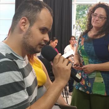 A imagem mostra o aluno Luis Gustavo Moreira de Andrade, do Ver com as Mos, sentado falando ao microfone. Em segundo plano est Mrcia Caspary, em p ao lado dele, o observando.