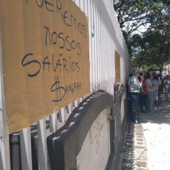 A foto mostra,  esquerda, um cartaz em papel pardo fixado na grade do IPC, com os dizeres (Texto) Queremos nossos salrios, Sanepar. (descrio) Ao fundo,  direita, h manifestantes em p.