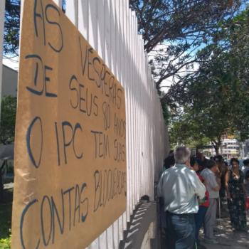 A foto mostra,  esquerda, um cartaz em papel pardo fixado na grade do IPC, com os dizeres (Texto) As vsperas de seus 80 anos, o IPC tem suas contas bloqueadas. (descrio) Ao fundo,  direita, h manifestantes em p.