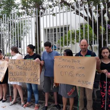 A foto mostra nove manifestantes em p, lado a lado, encostados na grade de entrada do IPC, segurando cartazes em papel pardo com os dizeres. (Texto) Sanepar, somos uma ONG, nos ajude. Cad a Justia do Paran? Bloqueou as contas do IPC. Sanepar, lucro lquido 2 semestre R$ 253,6 milhes.