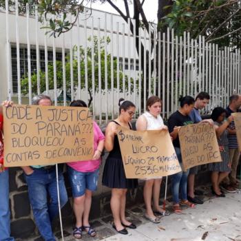 A foto mostra nove manifestantes em p, lado a lado, encostados na grade de entrada do IPC, segurando cartazes em papel pardo com os dizeres (texto) Cad a Justia do Paran? Bloqueou as contas de convnios. Sanepar. Lucro lquido 2 trimestre R$ 253,6 milhes. Cad a Justia do Paran? Bloqueou as contas do IPC.