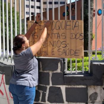 A foto mostra uma mulher de costas fixando na grade da entrada do IPC um cartaz em papel pardo com o texto Desbloqueia as contas dos convnios. Profs e sade.