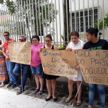 A foto mostra manifestantes em p lado a lado, encostados na grade do IPC, segurando cartazes em papel pardo com os dizeres (texto) Cad a Justia do Paran? Bloqueou as contas do IPC. Sanepar lucro lquido 2 trimestre 253,6 milhes.