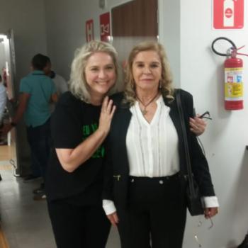 Professora Lilian em p, abraando por trs Mara Siaulys, da Fundao Laramara, que est  direita. Lilian passa o brao direito por trs de Mara e, com o esquerdo, a toca levemente no ombro. Elas sorriem. Lilian tem cabelo loiro at a altura do ombro. Usa camiseta preta e cala preta. Mara tambm tem cabelo loiro at o ombro, usa camisa de manga longa branca com casaco preto e cala preta.