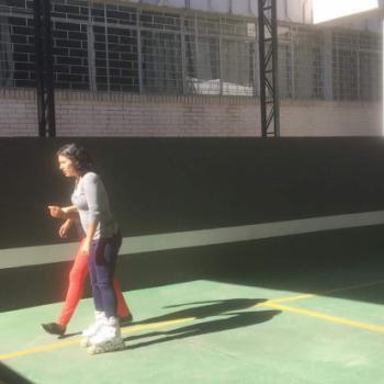 Uma aluna patinando na quadra. Ela est de lado para a foto, virada para a esquerda. Usa cala jeans e blusa cinza de manga comprida. Est concentrada no que faz.  direita dela, v-se apenas um brao a segurando e as pernas andando a seu lado.