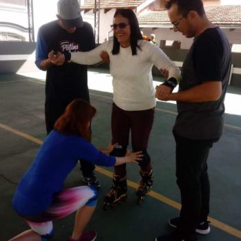 Uma mulher est sobre patins, olhando pra frente e sorrindo. Ela tem cabelo preto um pouco abaixo do ombro, usa blusa branca de manga longa e cala vermelha escuro.  apoiada pelos dois braos por dois jovens, que olham pra baixo.  esquerda, ele usa bon cinza, camiseta preta e cinza de manga longa e cala preta.  direita, ele tem cabelo preto e curto, usa culos de grau, camiseta cinza e preta e cala preta. Agachada em frente  mulher, est uma jovem de costas para a foto, tocando os joelhos da mulher sobre patins. Ela tem cabelos ruivos at o ombro, usa blusa azul de manga comprida e cala legging roxa, branca e azul.