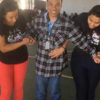 Um jovem est em p sobre patins. Ele sorri e olha pra frente. Tem cabelo preto, usa camiseta xadrez de preto e branco de manga longa e cala Jeans.  apoiado pelas duas mos por duas mulheres, que olham para baixo.  direita, ela tem cabelos pretos at o ombro, usa camiseta preta e cala vermelha.  direita, ela tem cabelos pretos e longos, usa camiseta preta e caa jeans.