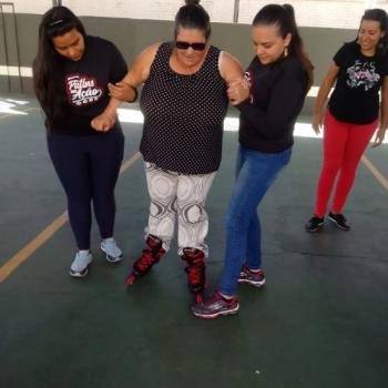 Uma mulher est em p sobre patins, olhando pra baixo. Tem cabelos pretos e presos, usa culos escuros, camiseta regata preta e cala branca e preta.  apoiada pelos dois braos por duas mulheres.  esquerda, a mulher tem cabelos longos e pretos, usa moletom preto e cala jeans.  direita, a mulher tambm tem cabelos longos e pretos, usa moletom preto e cala jeans. Em segundo plano,  direita, outra mulher assiste  cena. Ela tem cabelos pretos at o ombro, usa camiseta preta e cala vermelha.