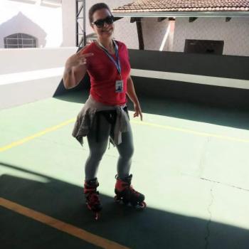 Uma mulher sorri posando para foto sobre patins, sorrindo e fazendo sinal com os dedos indicador e mdio abertos e os outros fechados. Ela tem cabelos presos, usa culos escuros, camiseta vermelha de manga curta, cala cinza e tem um moletom cinza amarrado  cintura.