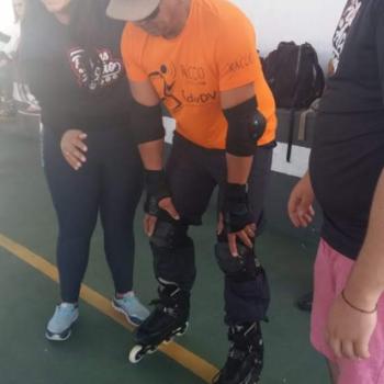 Um homem est em p sobre patins, com as costas levemente curvadas para baixo, apoiando as duas mos nos joelhos. Ele usa bon marrom, camiseta laranja e cala preta.  esquerda, uma mulher o segura e sorri olhando pra frente. Ela tem longos cabelos pretos, usa moletom preto e cala jeans.  direita, v-se parte de um homem em p, observando a cena. Ele tem cabelos pretos, usa culos de grau, camiseta preta e bermuda rosa.