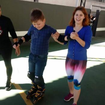 Um menino em p sobre patins olha para baixo e sorri. Ele tem cabelo castanho, usa camisa de manga longa azul e cala jeans. Do lado direito, uma mulher o segura pelo brao, olha para a foto e tambm sorri. Ela tem cabelos ruivos at o ombro, usa blusa azul de manga comprida e cala legging roxa, branca e azul.