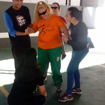 Uma mulher em p sobre patins sorri e olha pra baixo enquanto  apoiada pelos braos por uma mulher  direita, e um homem,  esquerda.  frente dela, h outra mulher agachada, de costas para a foto.