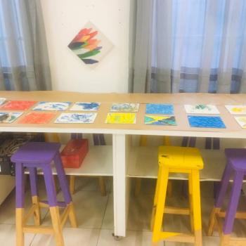 15 papis com pinturas esto expostos enfileirados sobre uma mesa retangular. Embaixo da mesa, h bancos coloridos.