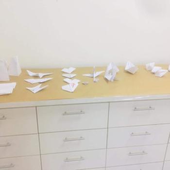 Dobraduras de barcos, aviões, pássaros e outros feitas em papel branco, expostas sobre um armário baixo com quatro filas de gavetas brancas.