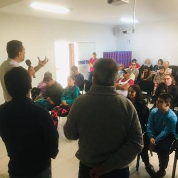 Em segundo plano, de costas para a foto e de frente para o pblico, Enio Verri fala segurando o microfone com a mo direita e gesticulando com a esquerda. As pessoas esto sentadas em cadeiras, srias e atentas. H crianas, jovens e adultos. Em primeiro plano, de costas, dois homens em p lado a lado.