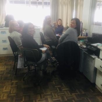 Imagem de seis mulheres sentadas em grupo, olhando para trás e sorrindo para a foto. Elas estão numa sala de escritório, com móveis brancos no entorno delas.