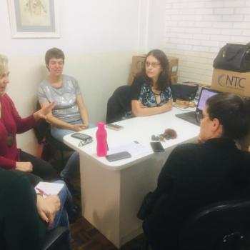 Imagem de cinco pessoas sentadas em roda em volta de uma mesa.  direita, v-se somente o brao de uma sexta pessoa.  esquerda, uma mulher de cabelos loiros e blusa roxa fala, gesticulando a mo esquerda. As outras pessoas prestam ateno nela.