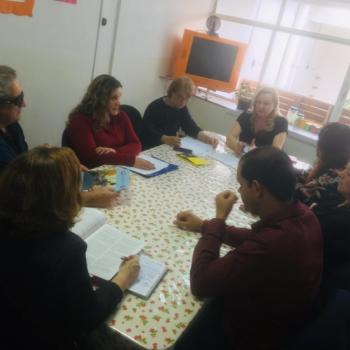 Imagem de oito pessoas sentadas em volta de uma mesa retangular, conversando. Elas esto com papis sobre a mesa, algumas escrevendo.