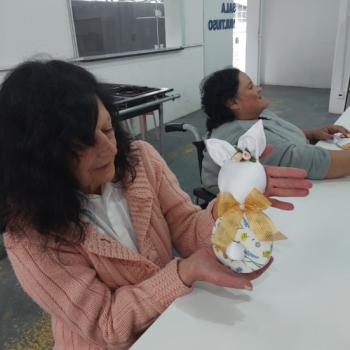 Duas mulheres sentadas em uma mesa branca dentro de uma sala clara e ampla. A mulher em primeiro plano, de cabelos escuros e cacheados, veste um casaco rosa claro e segura nas mos um artesanato em formato de coelho, feito com tecido estampado, lao e orelhas grandes. A outra mulher ao fundo est em uma cadeira de rodas e sorri, parecendo observar a cena ou conversar. Ao fundo, v-se uma porta de vidro com a inscrio SALA DE MULTIUSO e uma janela grande. O ambiente transmite sensao de criatividade e convivncia.