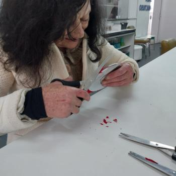 Foto de duas mulheres sentadas ao redor de uma mesa branca. A mulher em primeiro plano tem cabelo preto e veste um casaco claro; ela segura uma rosa vermelha feita de papel, com caule verde. Ao lado dela, outra mulher est falando ao celular, segurando o aparelho vermelho na orelha e usando culos escuros. H tecido amarelo e alguns objetos em cima da mesa, sugerindo talvez uma oficina de artesanato.