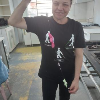Mulher em p, sorrindo, dentro de uma cozinha com armrios e fogo ao fundo. Ela tem cabelo preso e usa uma camiseta preta com smbolos de pessoas guiando outras; nas palavras esto escrito 