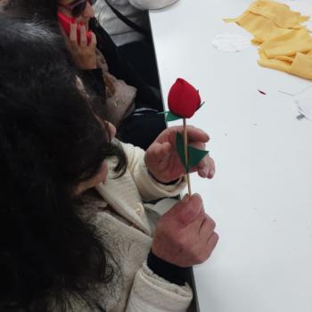 Senhora de cabelos escuros sentada  mesa branca, concentrada ao cortar um papel vermelho com uma tesoura. Ela veste um casaco bege claro e h pedaos pequenos de papel vermelho sobre a mesa. Ao fundo,  possvel ver prateleiras com objetos, cadeiras e uma porta com o letreiro SALA MULTIUSO. Tambm h outra tesoura sobre a mesa. A iluminao do ambiente  clara e parece aconchegante.