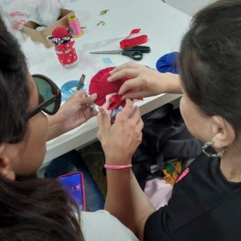 Duas pessoas esto sentadas lado a lado, trabalhando juntas em um artesanato colorido com tecido vermelho e fitas. Sobre a mesa h materiais como tesoura, linha, cola quente e enfeites usados na atividade.