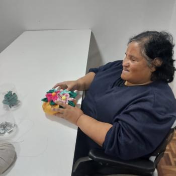 Uma mulher est sentada em uma cadeira de rodas, sorridente, enquanto segura um arranjo artesanal colorido feito de tecido, com flores em tons de rosa, amarelo, vermelho e verde. Ela est em uma sala simples, com uma mesa branca ao lado e algumas peas de tecido ou materiais de artesanato sobre a mesa. A mulher veste uma blusa escura e tem cabelos escuros ondulados. A atmosfera transmite acolhimento, foco e bem-estar durante a atividade manual.