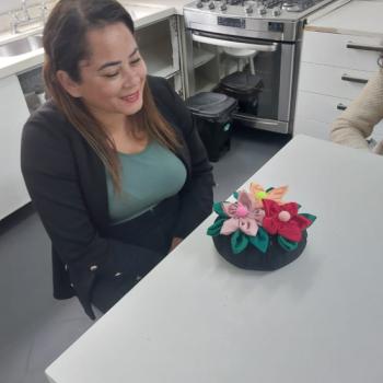 Mulher sentada  mesa em uma cozinha moderna, olhando sorridente para um arranjo de flores de tecido coloridas sobre a mesa. Ao fundo, h um fogo prateado com forno embutido, prateleiras abertas e uma pia  esquerda. A mulher tem cabelos castanhos e veste um blazer preto com blusa cinza. A cozinha est bem iluminada e com aparncia organizada.