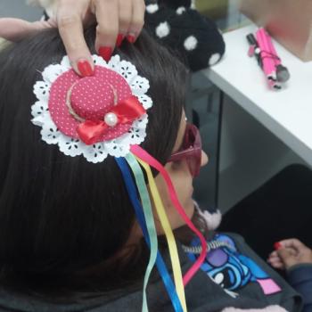 Uma pessoa segura na cabea um enfeite artesanal feito com tecido vermelho de bolinhas, lao de fita, prola e fitas coloridas penduradas. O adorno lembra um pequeno chapu decorativo, smbolo da atividade de artesanato.